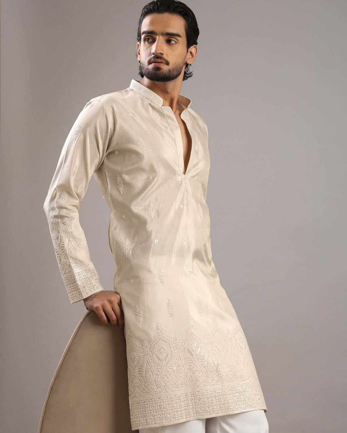 A man modeling a cream-colored, long-sleeved kurta with subtle embroidery and a mandarin collar, paired with white trousers, against a solid gray studio background.