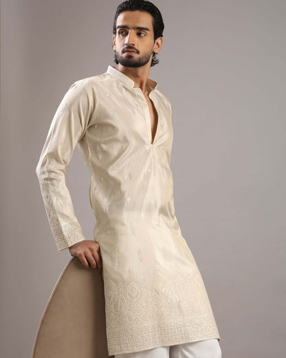 A man modeling a cream-colored, long-sleeved kurta with subtle embroidery and a mandarin collar, paired with white trousers, against a solid gray studio background.